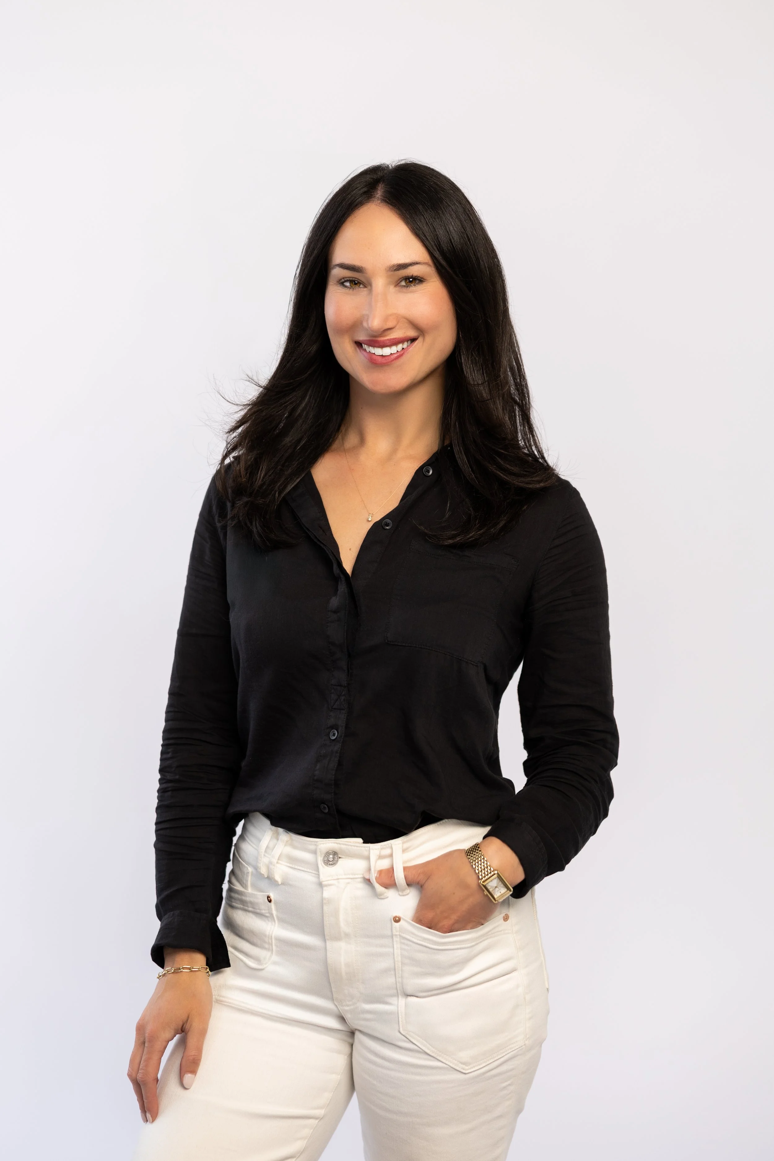 A woman with long dark hair, wearing a black button-up shirt and light-colored pants, smiling in front of a plain white background.