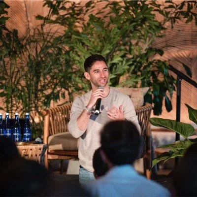 A man holding a microphone speaking at an indoor event with plants and seating in the background