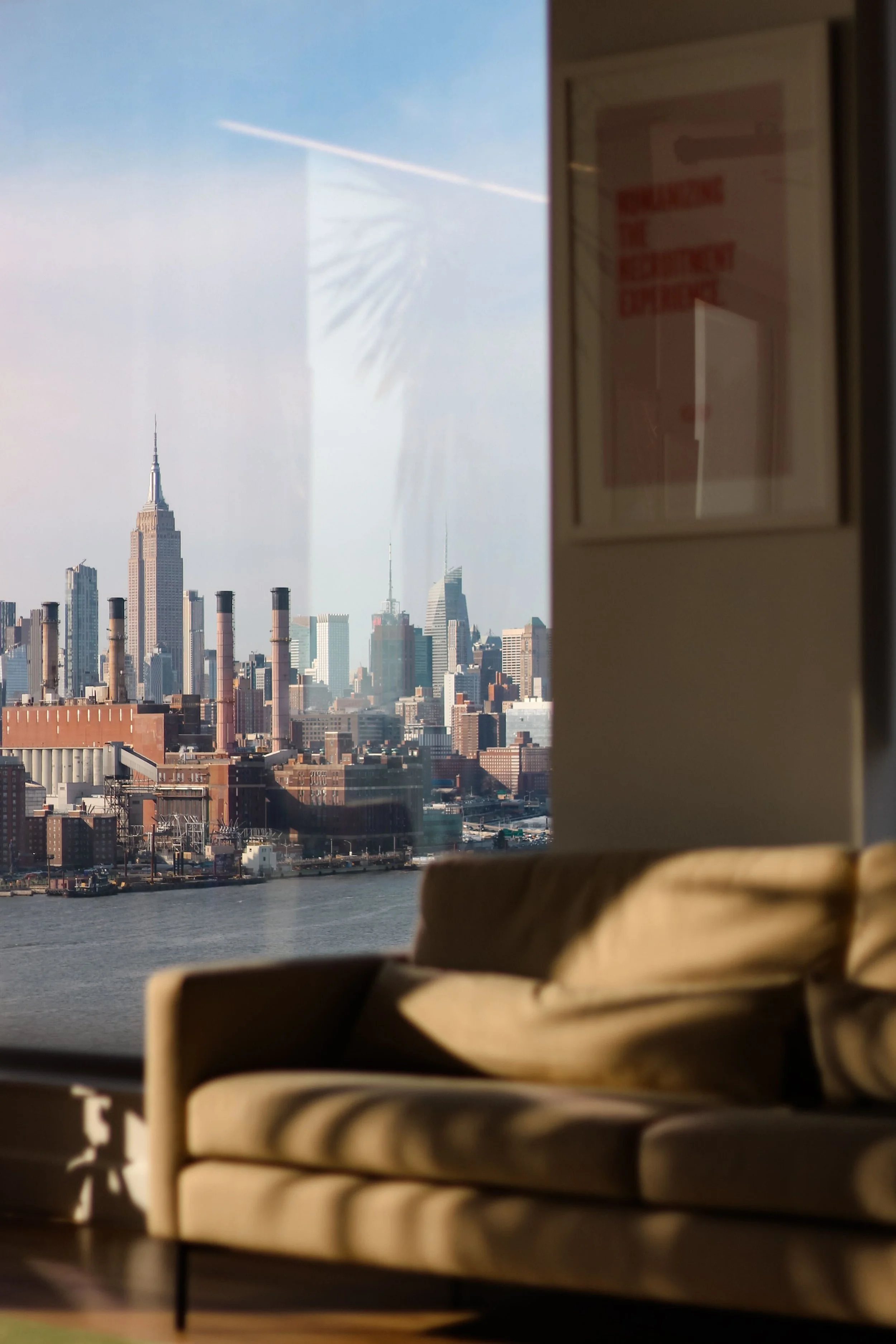 View of the New York City skyline through a window, featuring the Empire State Building and other skyscrapers, with a beige couch and shadowed interior in the foreground.