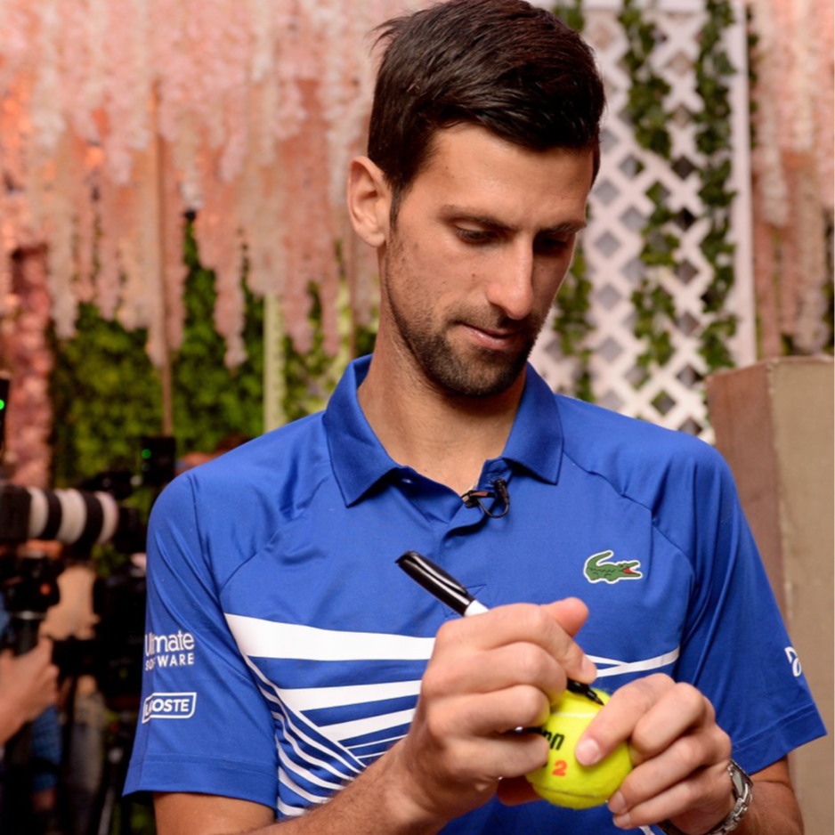 A man with dark hair and a beard wearing a blue Lacoste sports shirt is signing a tennis ball with a black marker, in a setting with a pink floral backdrop and some greenery.
