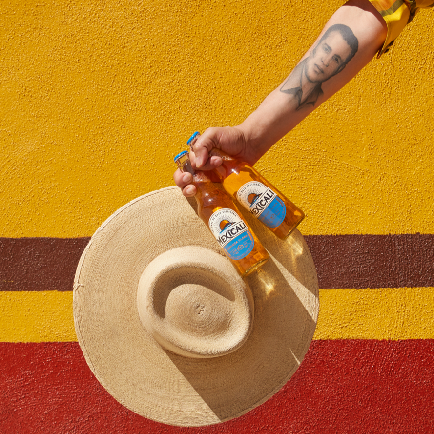 A person holding two bottles of Mexican beer against a yellow wall with red and brown stripes. A straw hat is placed on the ground.