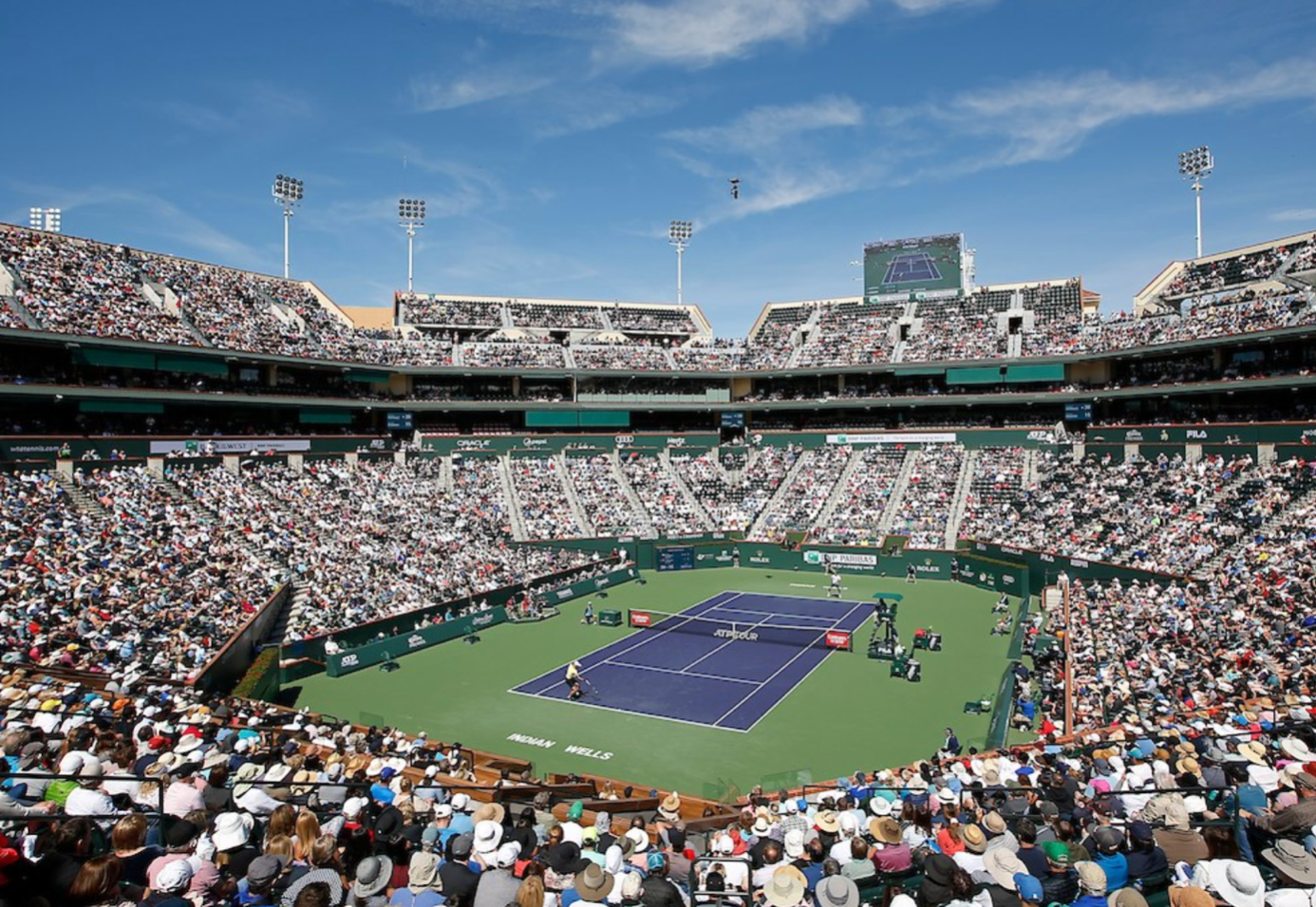 A crowded tennis stadium with players on a blue court under a clear blue sky, filled with spectators in wide-brimmed hats and caps.
