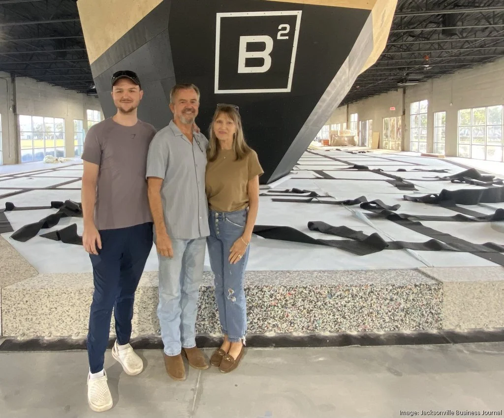 Three people standing inside a large climbing gym with a bouldering wall that has a sign labeled ‘B2’. The background shows indoor climbing terrain with padded flooring and large windows.