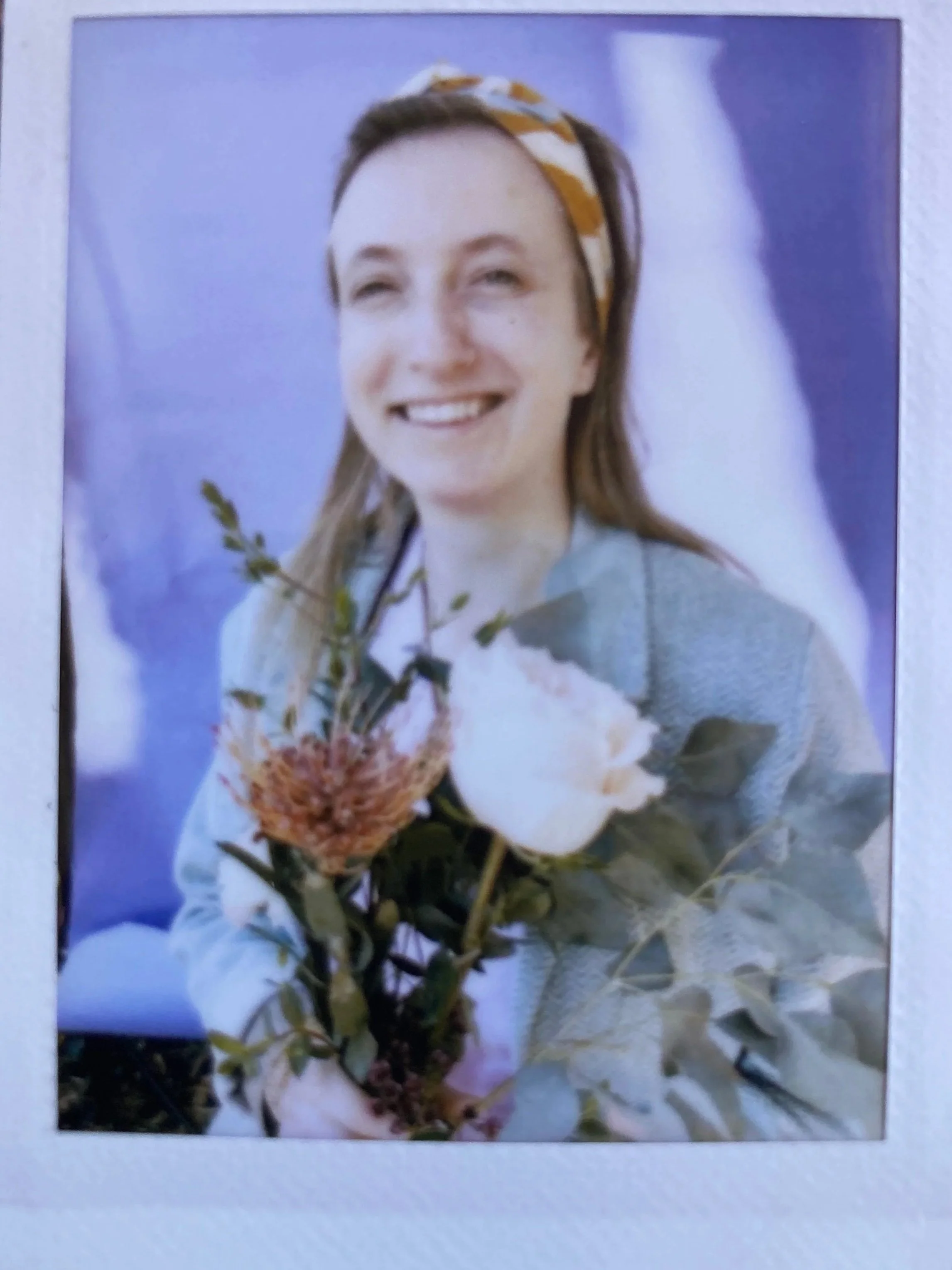 A mid-thirties woman smiling while holding a bouquet of flowers, wearing a yellow patterned headband and a gray blazer against a purple background.