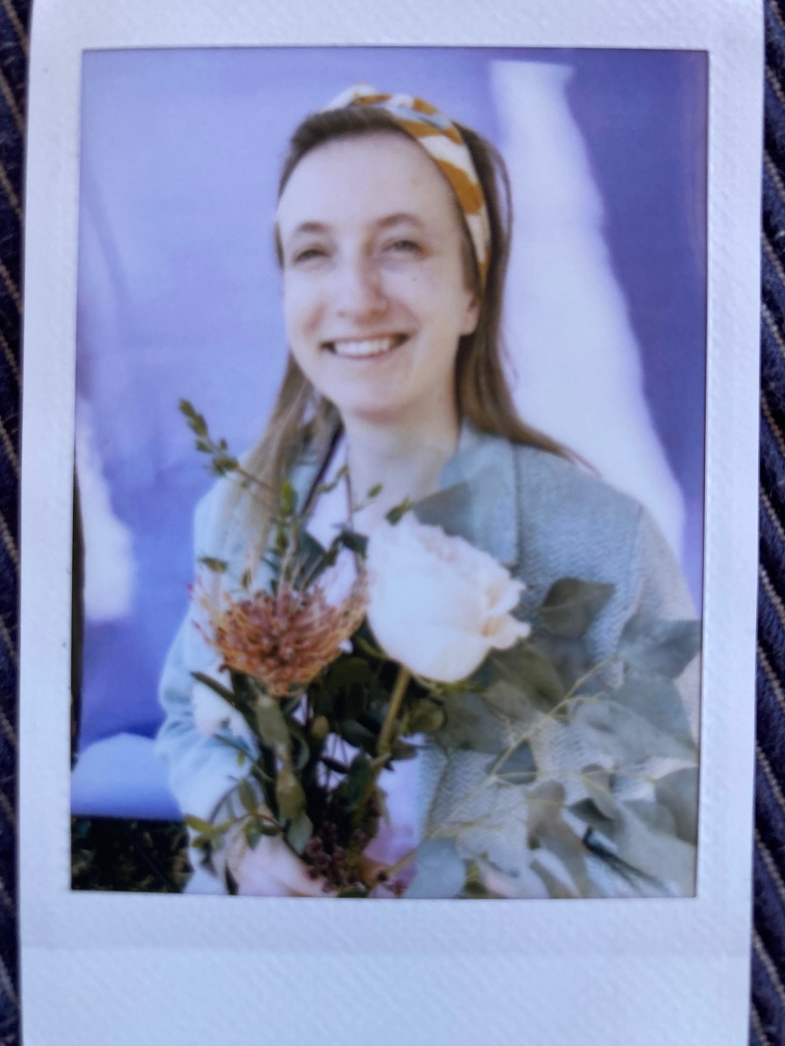A mid-thirties woman smiling, wearing a headband, holding a bouquet of flowers including a white rose and greenery, with a purple background.