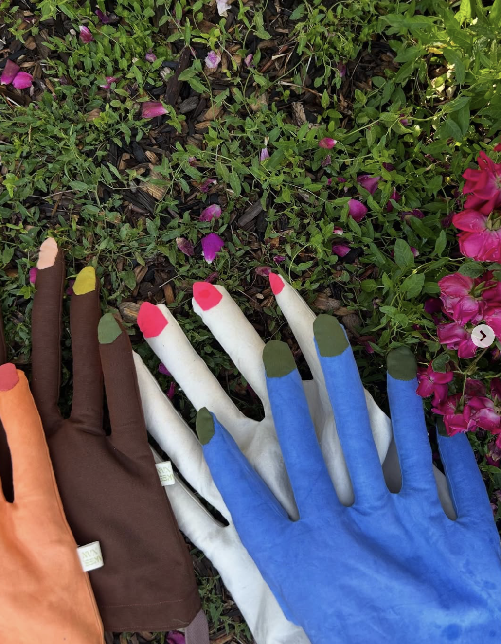 Five colorful hand bags placed on the ground with green and purple plants and flowers in the background.