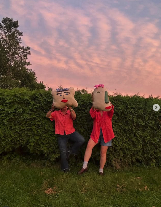 Two people standing outdoors at sunset, wearing large plush head masks resembling human faces, dressed in red shirts and standing in front of a hedge.