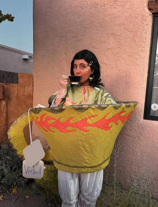 Person dressed in a tea cup costume with flames, holding a tea cup, standing outside near a pink wall.