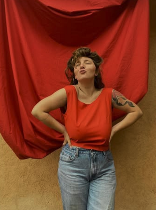 Woman with short curly hair wearing a red sleeveless top and jeans, standing in front of a red cloth backdrop with her hands on her hips, making a puckered lip expression.
