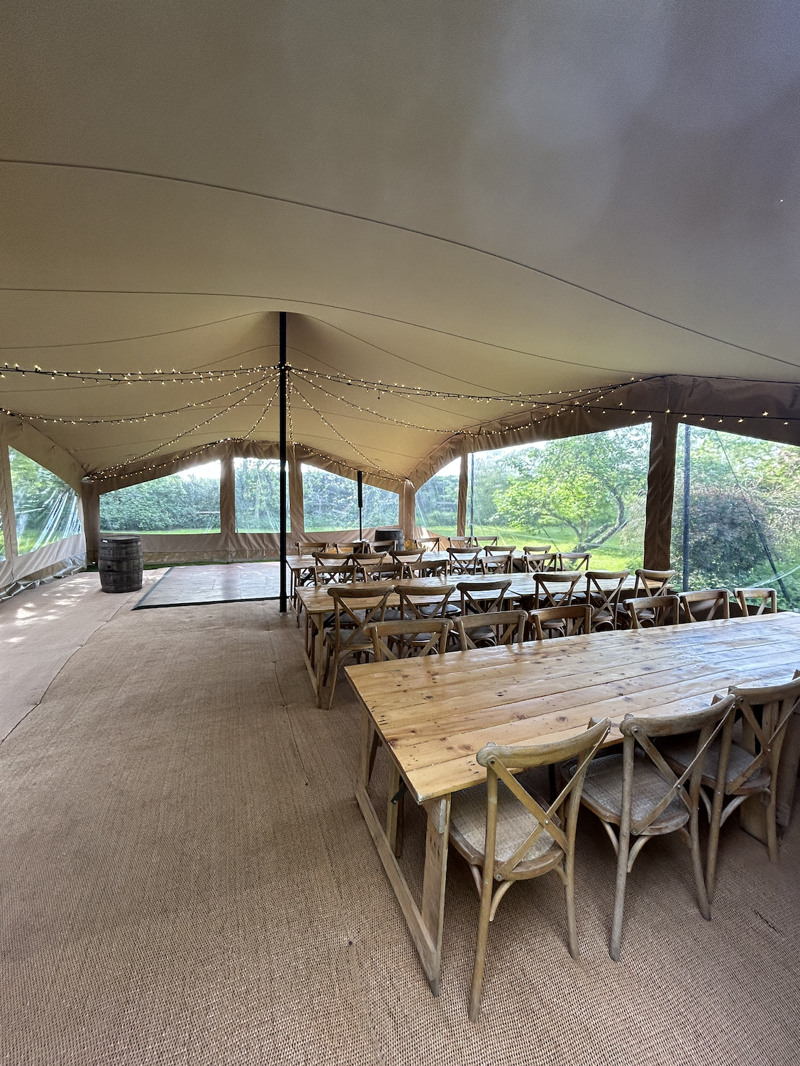Empty outdoor tent with wooden tables and chairs, string lights hanging from the ceiling, and a view of green trees outside.
