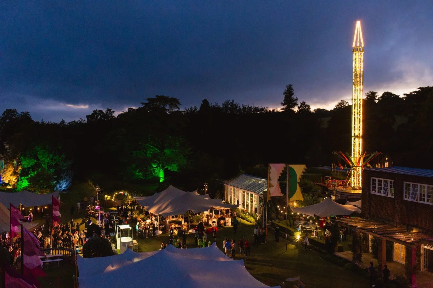 Night scene of an outdoor fair or festival with tents, colorful lighting, and a tall illuminated amusement ride in the background, surrounded by trees and a dark, cloudy sky.