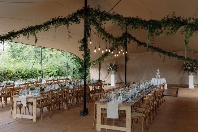 Long wooden banquet tables set with glassware, plates, and silverware inside a large tent with hanging string lights and greenery, prepared for a wedding or event.