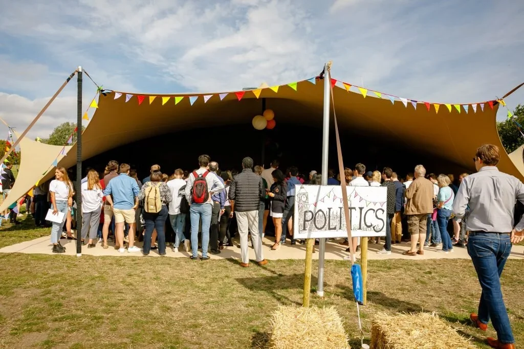 People gathered under a large canopy tent decorated with colorful pennant banners at an outdoor event, with a sign labeled 'Politics' in front.