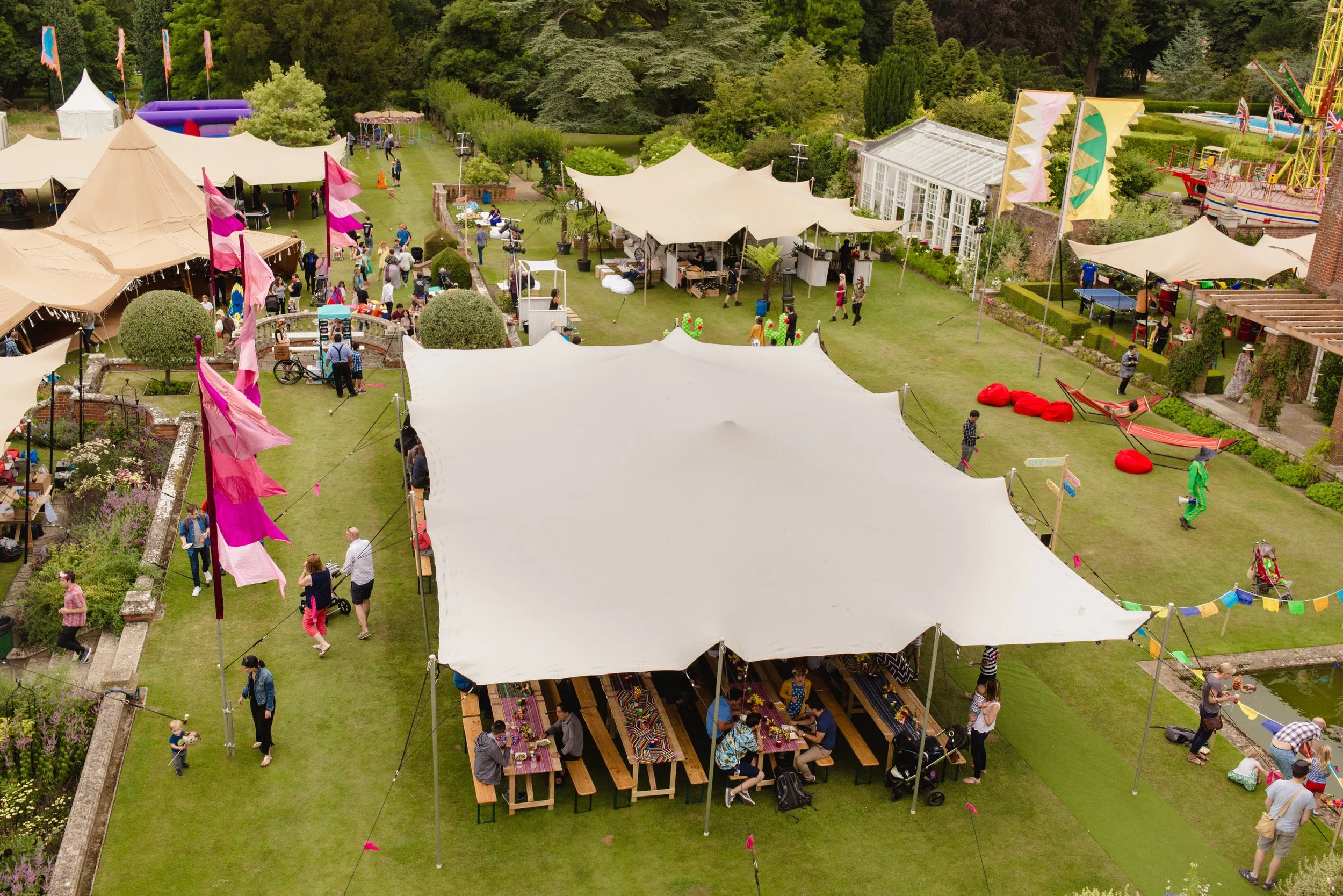 Aerial view of a lively outdoor fair or festival with white tents, flags, people walking, sitting at picnic tables, and children playing on a grassy area with trees.