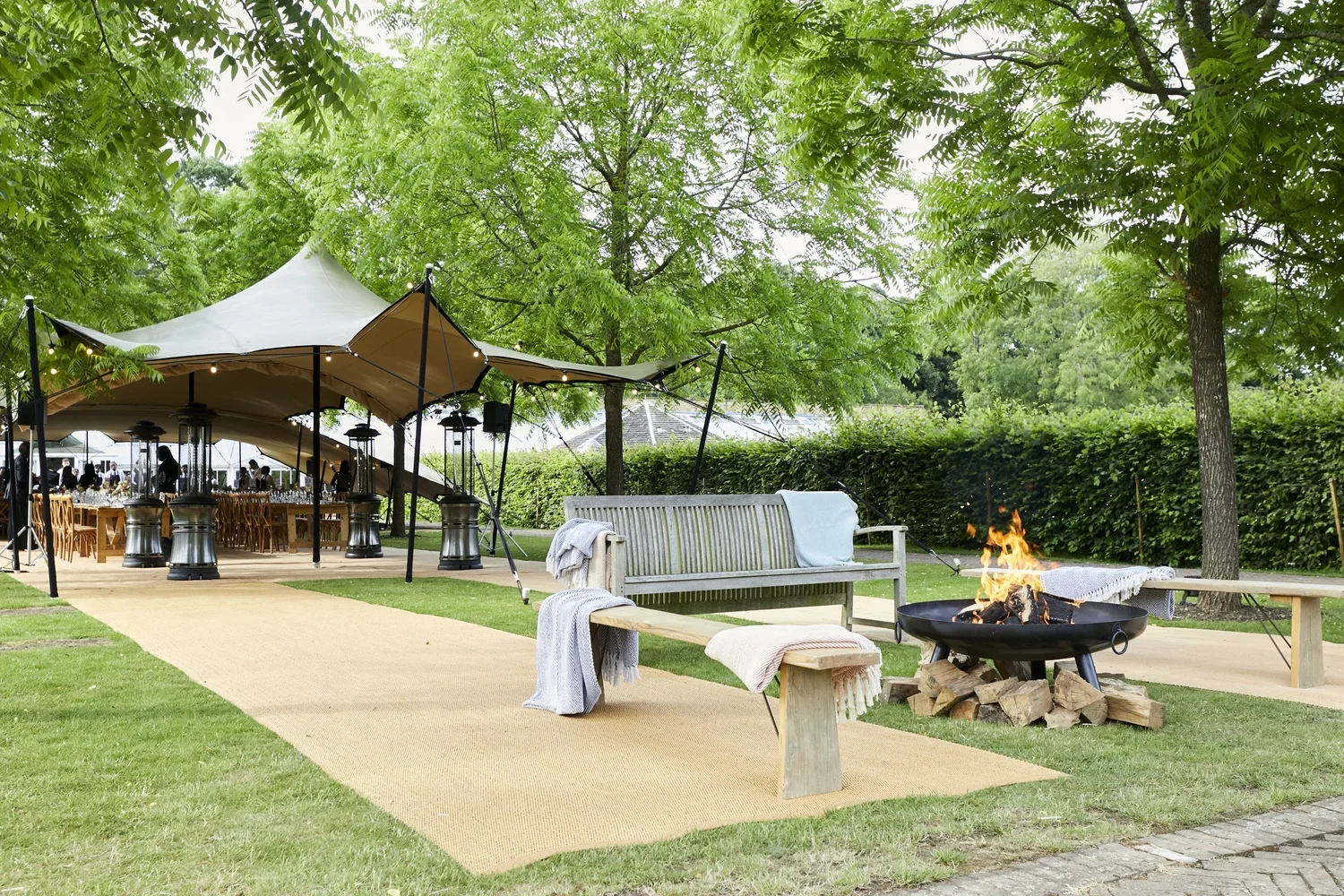 Outdoor event setup with a large canopy tent, tables, and chairs, surrounded by trees and greenery, with a fire pit and benches on a grassy area.