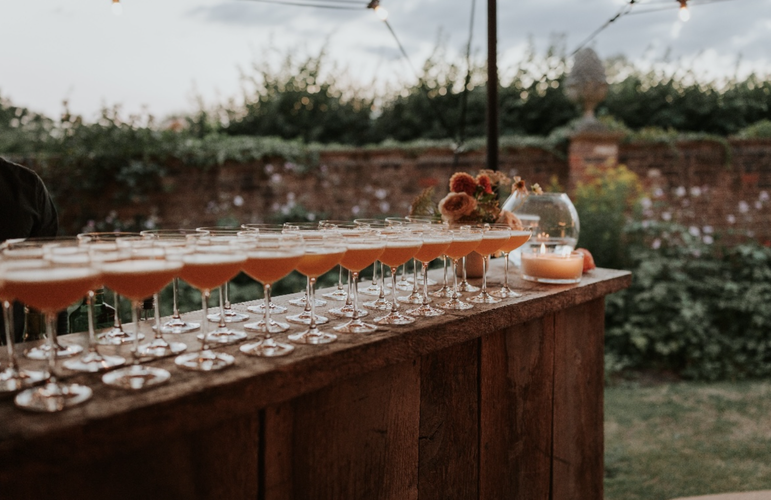 A bar table outdoors with numerous pink cocktails in glasses, decorated with flowers, set on a wooden surface at dusk with a garden and brick wall in the background.