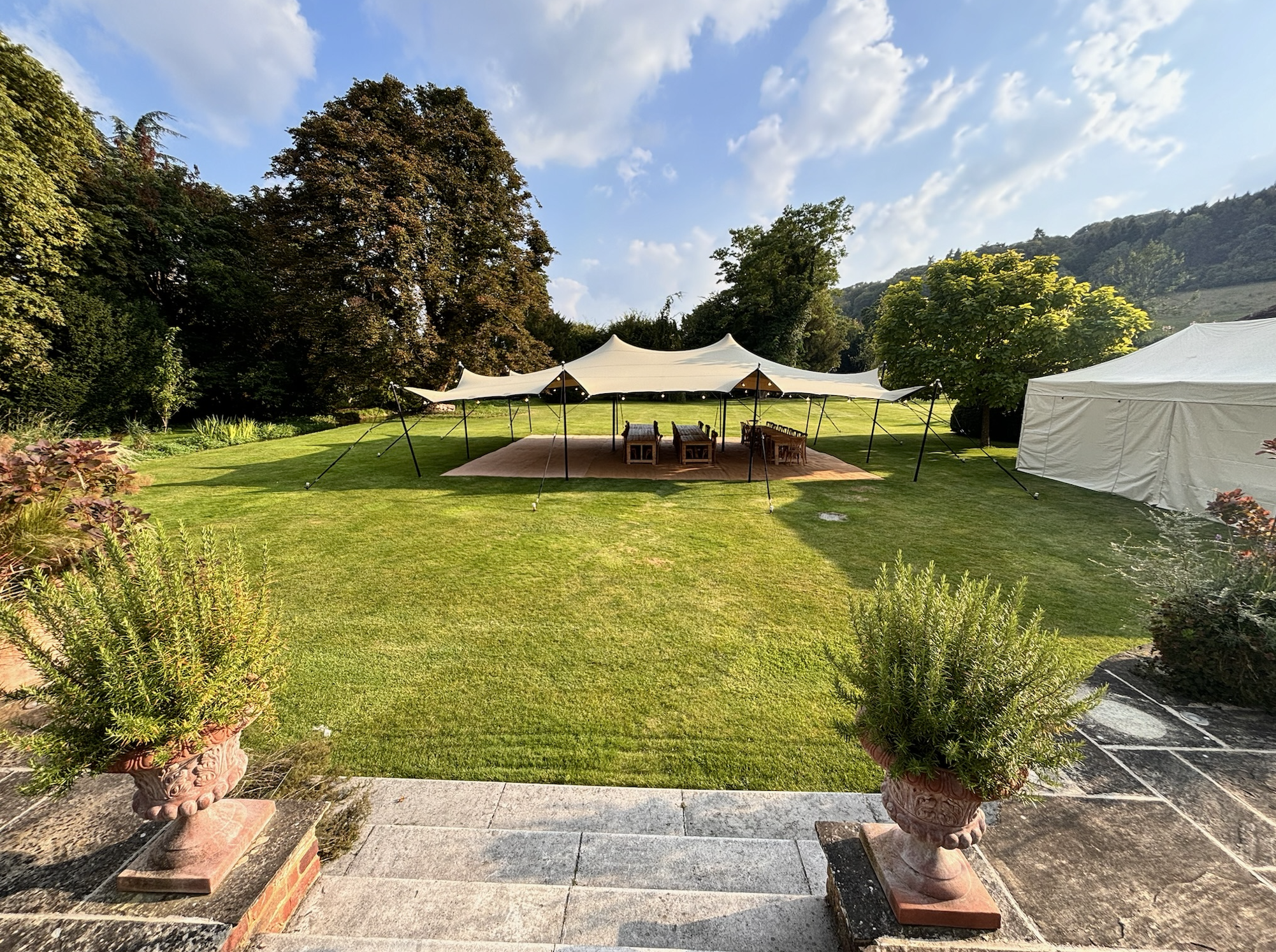 A spacious outdoor garden area with a large white canopy tent, chairs, and a lawn surrounded by trees under a partly cloudy sky.