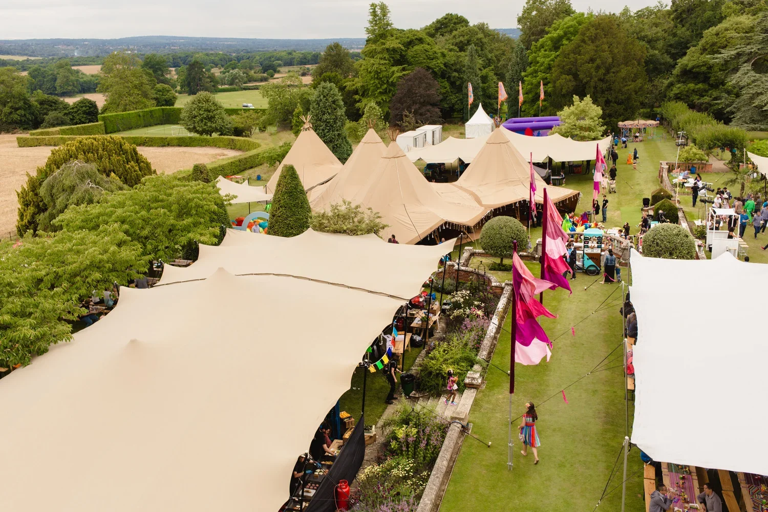 Outdoor market or festival with beige tents, flags, and people walking along a grassy path surrounded by trees and colorful flowers.