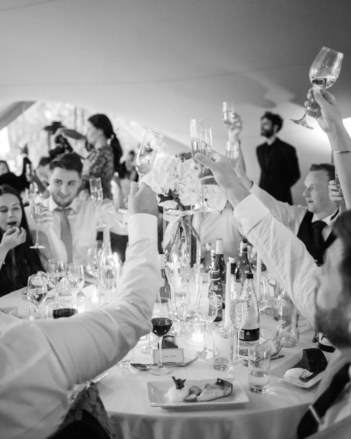 Elegant dinner party inside a large tent with decorated tables, flowers, candles, and string lights, with people socializing and a band performing in the background.