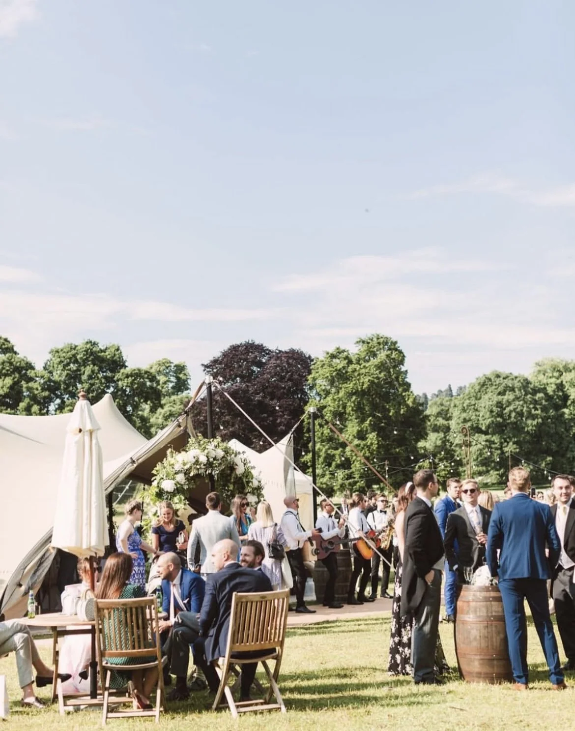 People gathered at an outdoor event under a clear sky with large trees in the background. Some are sitting at tables, while others are standing and drinking. There is a tent with floral decorations and a group of musicians playing guitars.