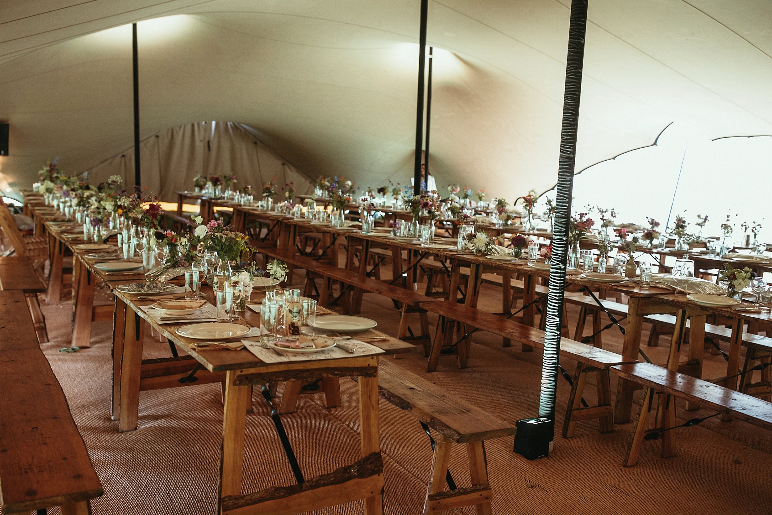 Outdoor event under a large tent with long wooden tables decorated with colorful flowers and small candles, set up for a celebration or gathering on a grassy area.