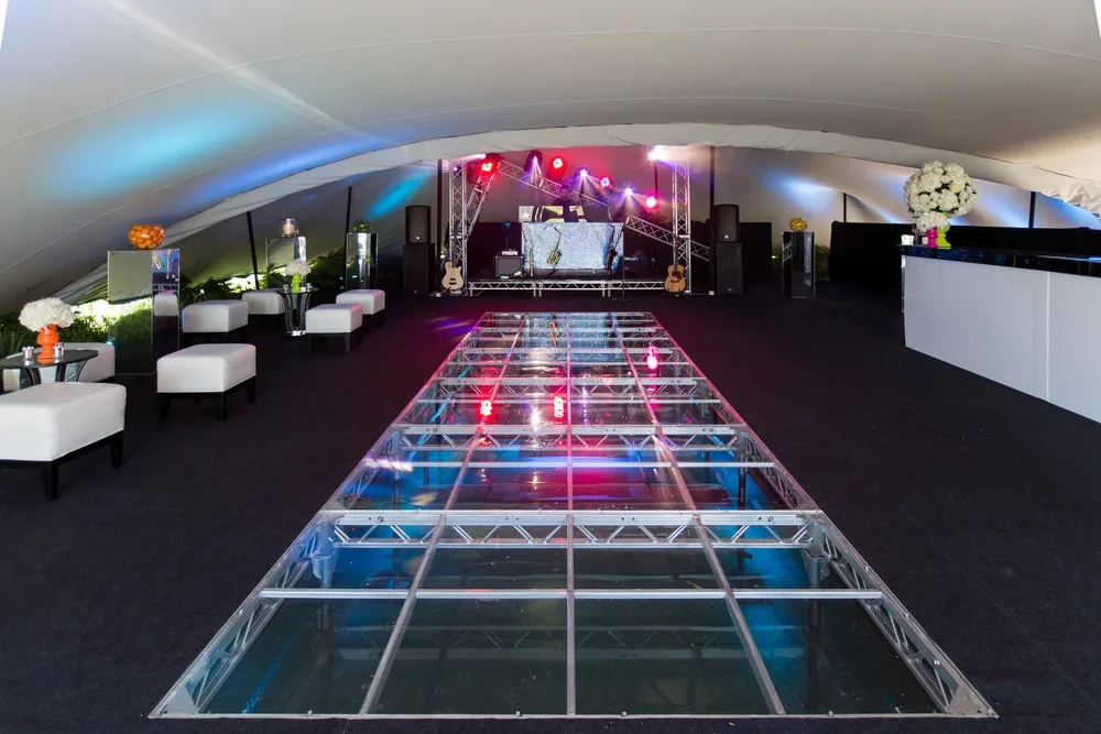 Empty event space with a glass dance floor, stage with musical instruments, DJ equipment, and colorful lighting.