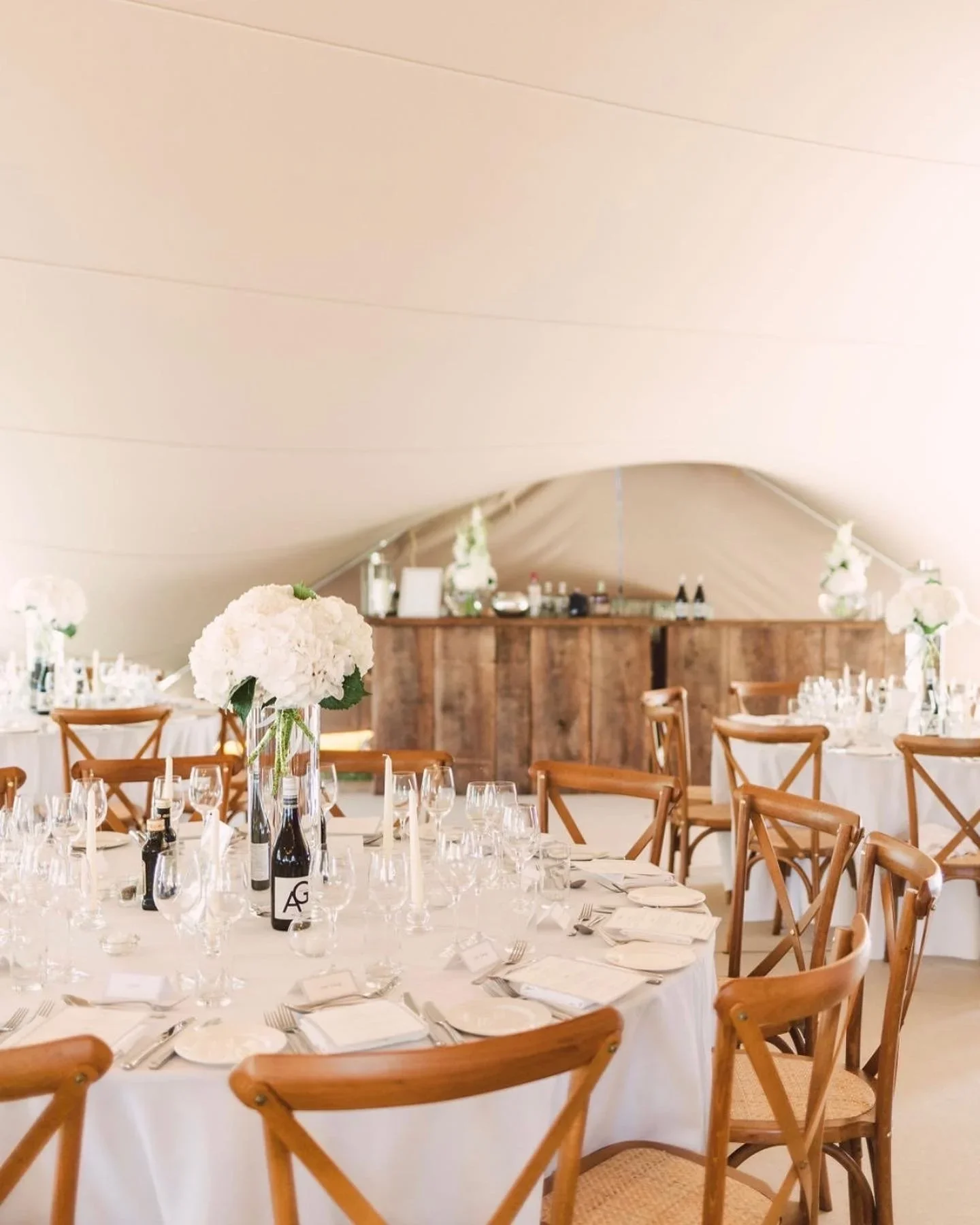 Elegant wedding reception under a large white tent with round tables decorated with white floral centerpieces and set with glassware, plates, and silverware, and wooden chairs surrounding the tables.