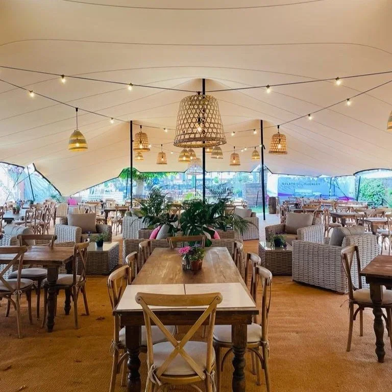 Interior of a bright restaurant with wooden tables, wicker chairs, and greenery. Hanging lamps and string lights decorate the ceiling, with large windows allowing natural light.