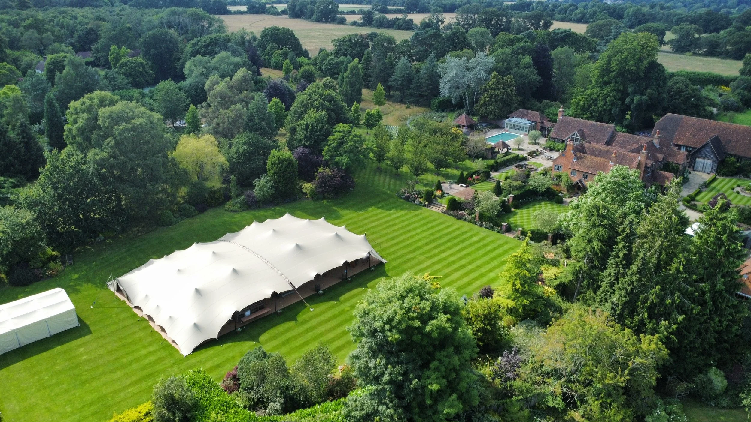 Aerial view of a spacious backyard with a large white tent, manicured lawn, lush trees, and a mansion with a swimming pool in the distance, surrounded by countryside.