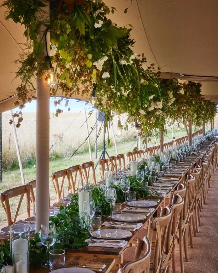 Long dining table set with plates, wine glasses, and silverware, decorated with white candles and lush green foliage, under a canopy with hanging greenery and string lights in a rustic outdoor setting.