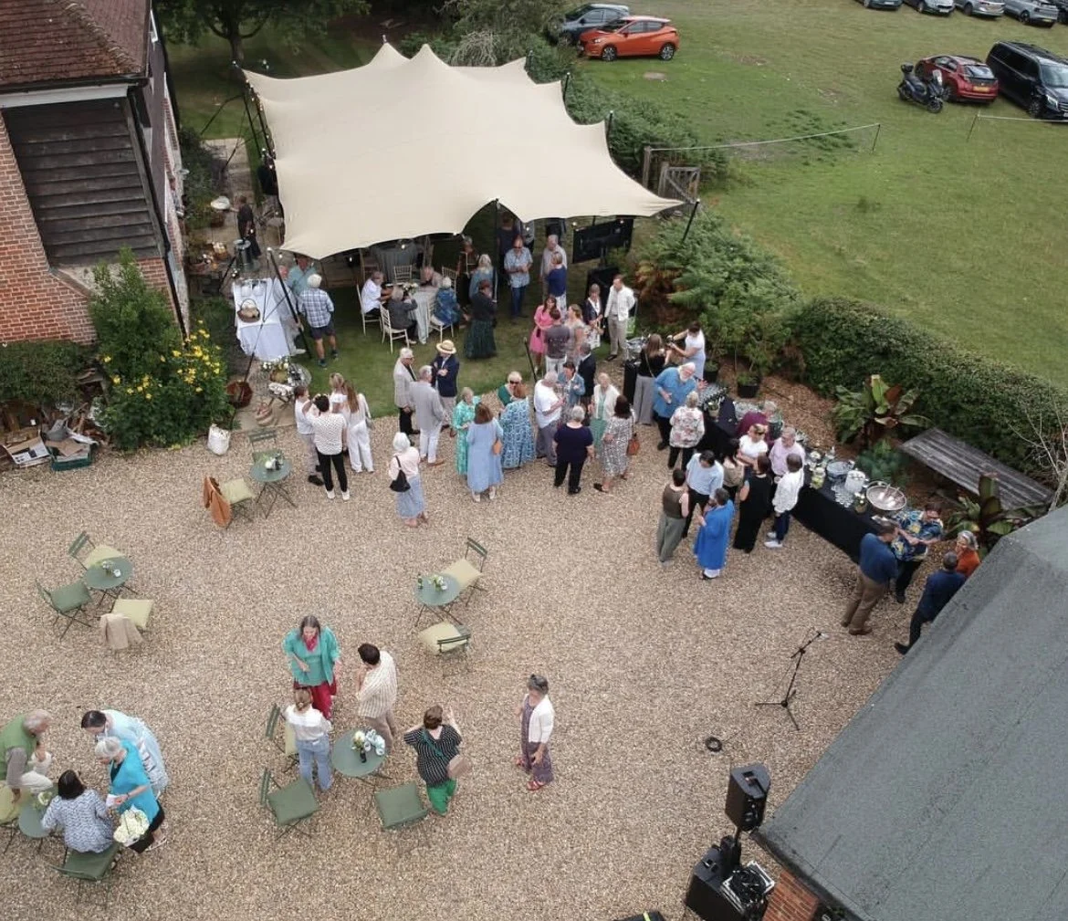 People gathered at a backyard outdoor party with a white canopy, dining tables, and seating area, on a gravel patio with green grass and parked cars in the background.