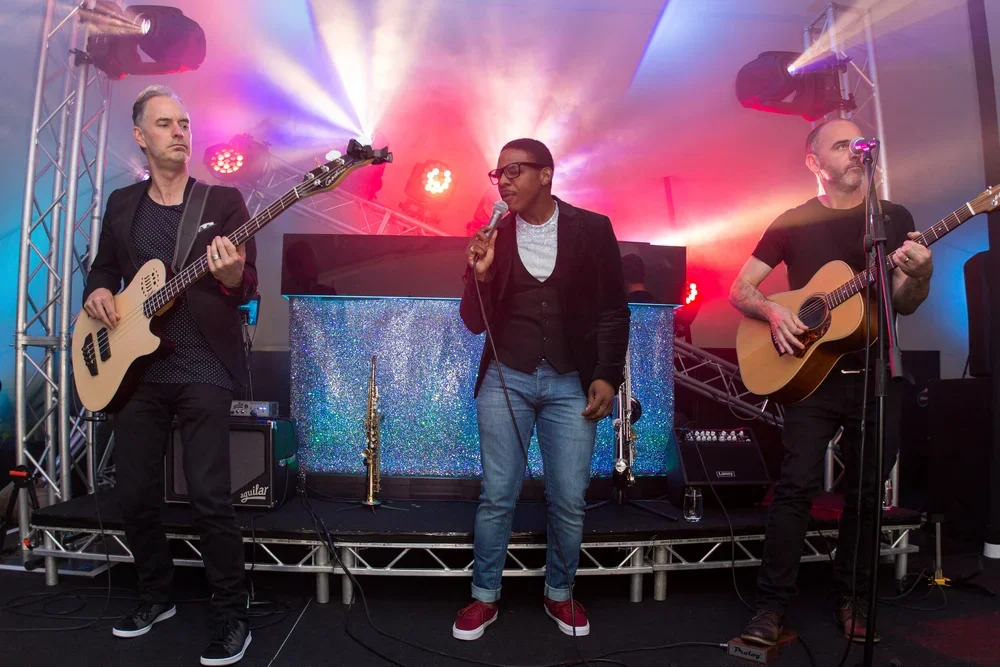 Three musicians performing on stage with colorful lights, one woman singing into a microphone, one man playing bass, and another man playing acoustic guitar.