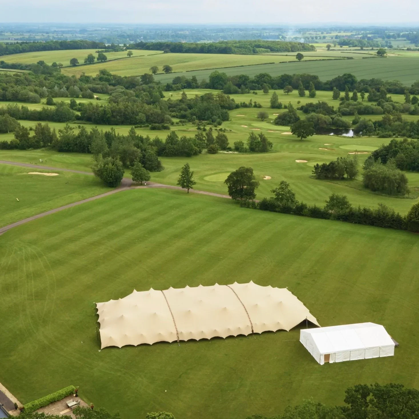 A large beige event tent and a smaller white tent set up on a grassy area of a golf course surrounded by trees and rolling green hills.