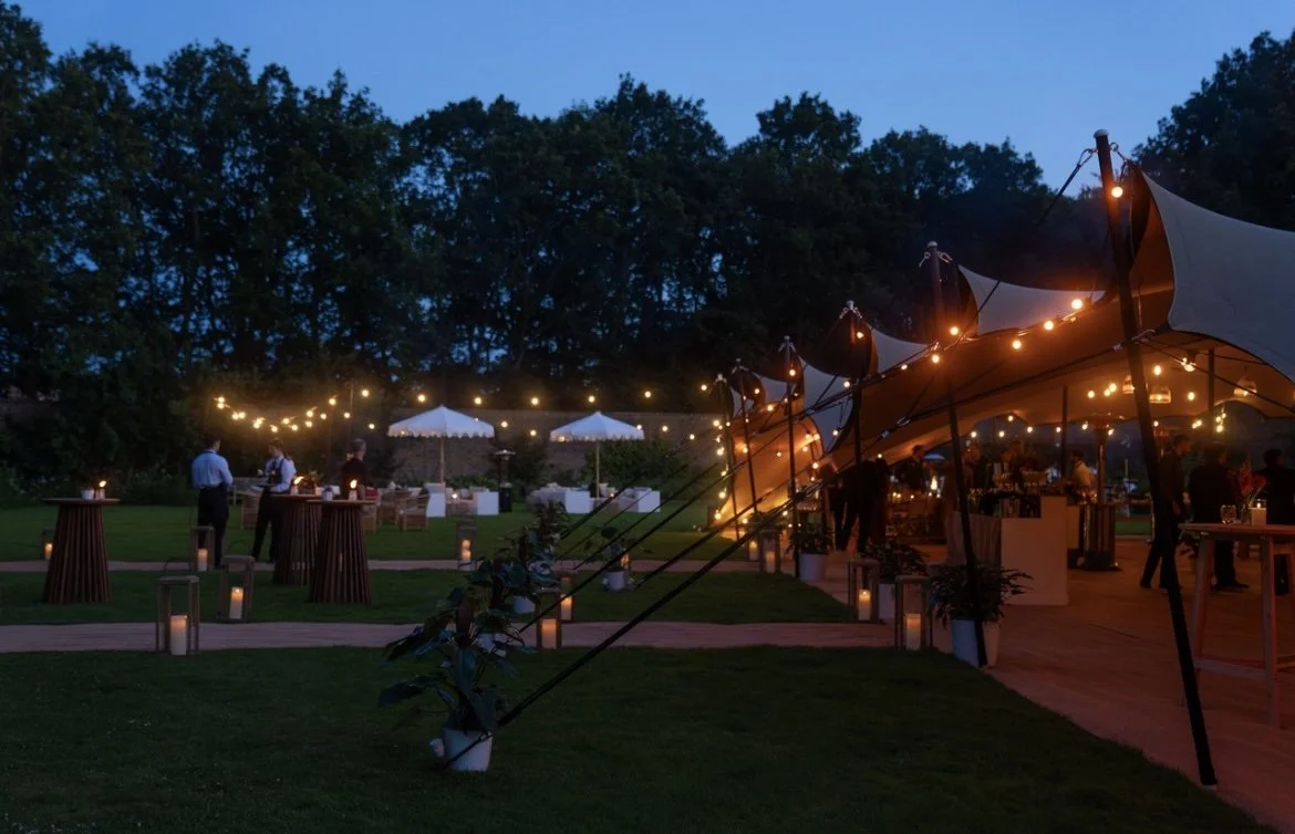 A large outdoor event tent with multiple peaks set up on a grassy lawn, with a smaller tent in front. The sky is cloudy and the area is cordoned off with chains and wooden posts.