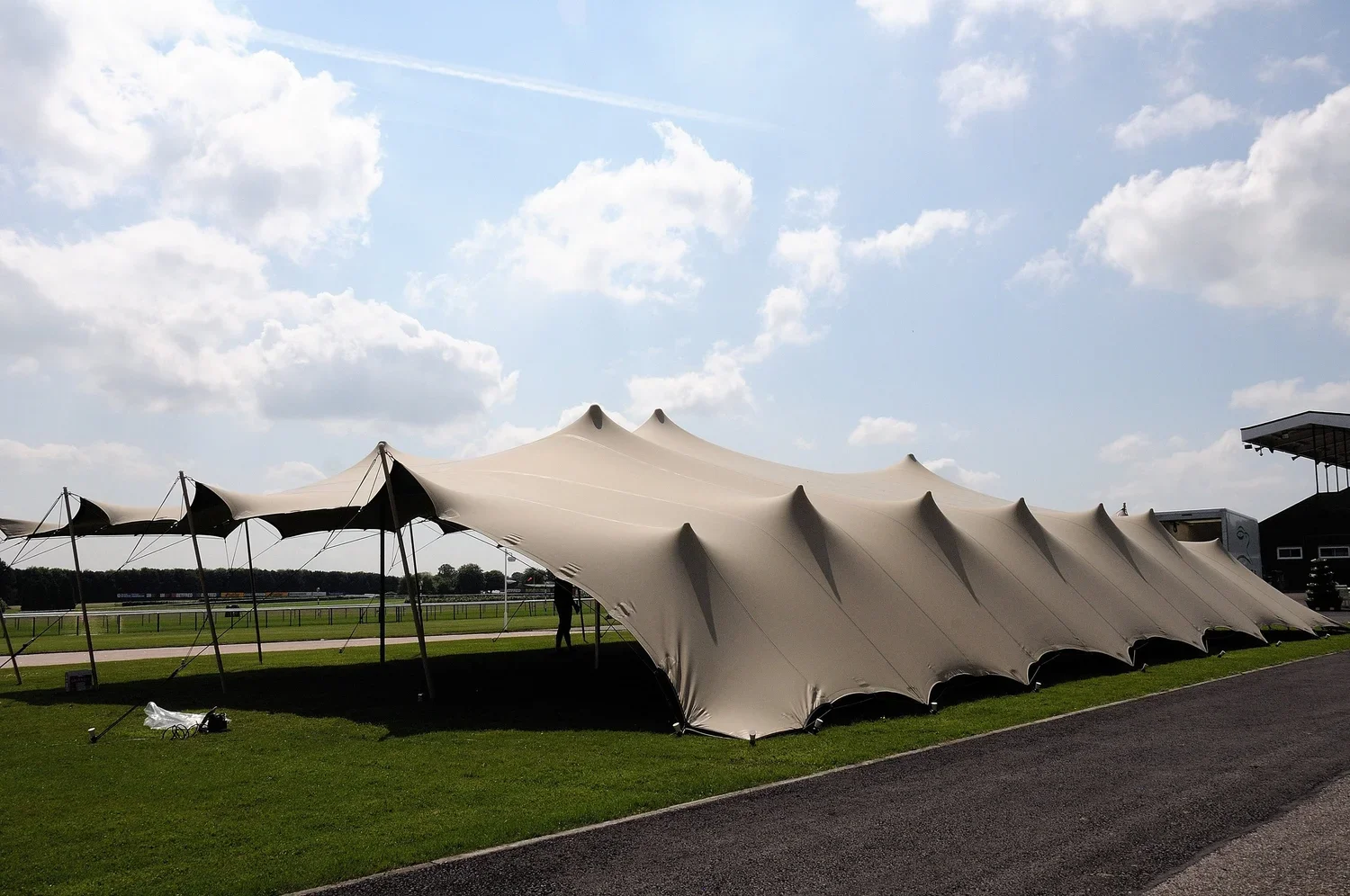 Large white tent with peaks set up on a grassy area at what appears to be a racetrack, under a partly cloudy sky.
