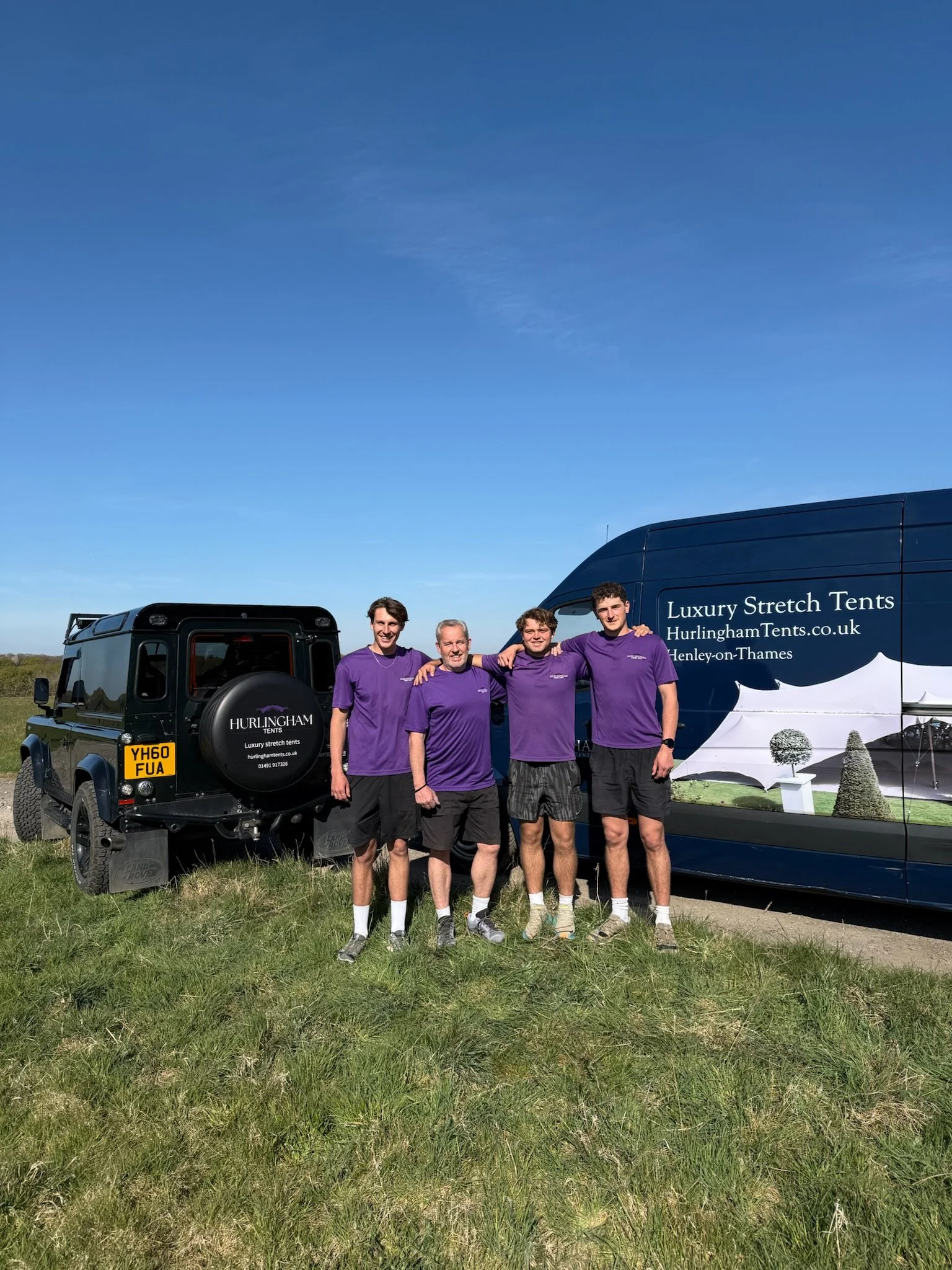 Four men in purple shirts standing outdoors on grass, smiling and posing with their arms around each other, with two vehicles, one black and one blue, advertising luxury stretch tents.