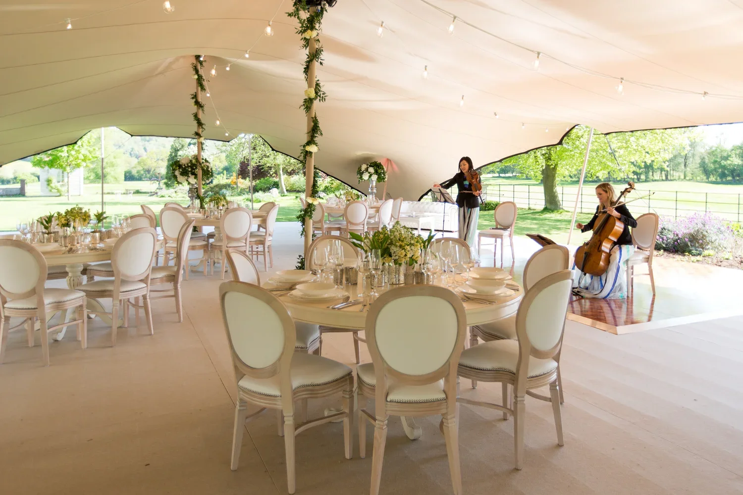 Elegant outdoor event tent with round dining tables, white chairs, floral centerpieces, and live musicians playing in a lush green park setting.