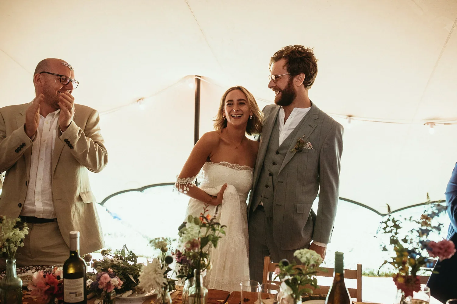A bride and groom sharing a joyful moment at their wedding reception, surrounded by friends and decorated with flowers and bottles on the table.