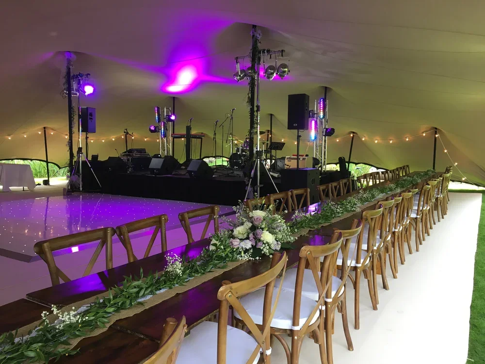 Long banquet table decorated with greenery and flowers, set under a tent with a stage and musical equipment, purple lighting, and string lights.