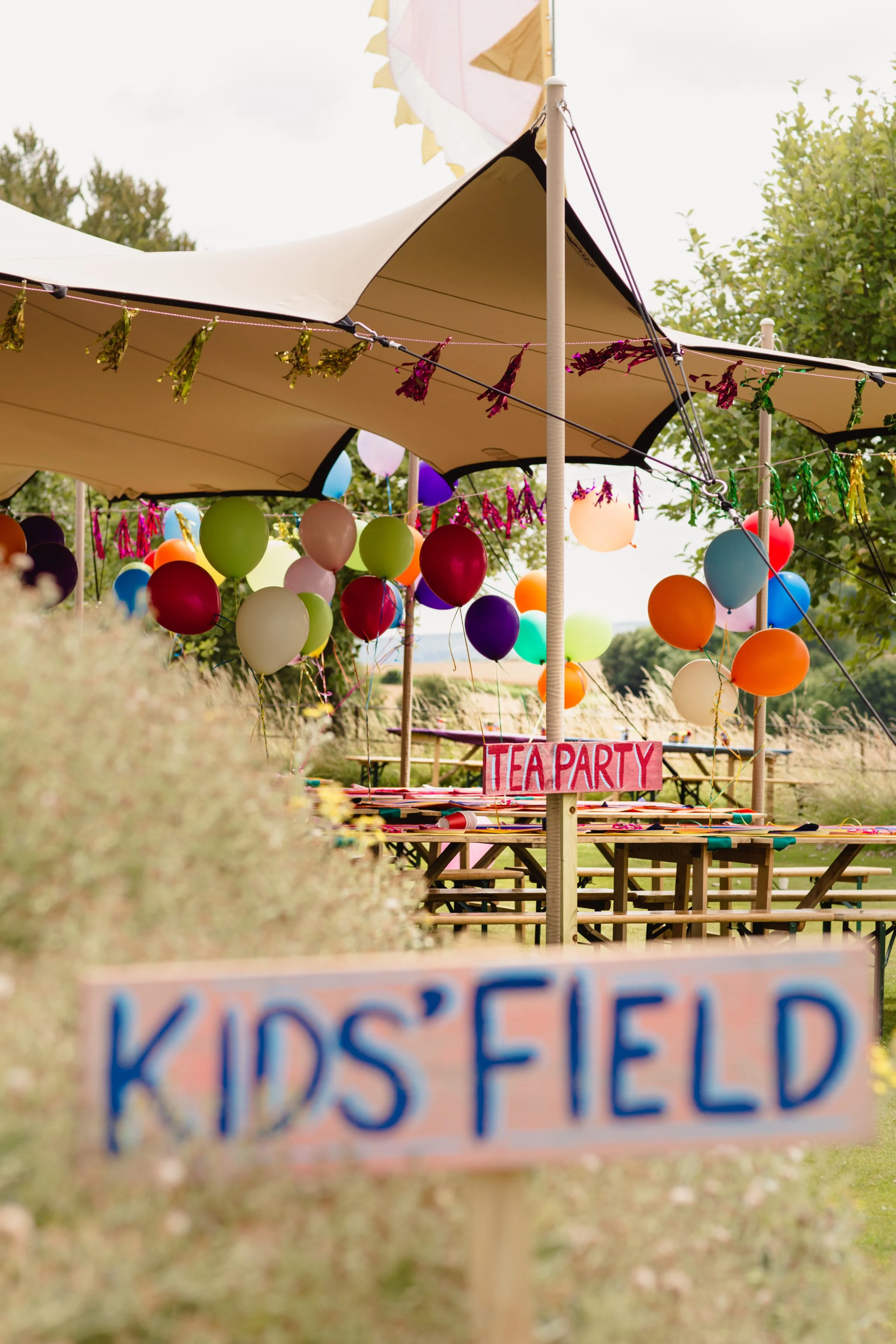 Outdoor kids' party area with colorful balloons, balloons, and signs reading 'Tea Party' and 'Kids' Field' under a large canopy.