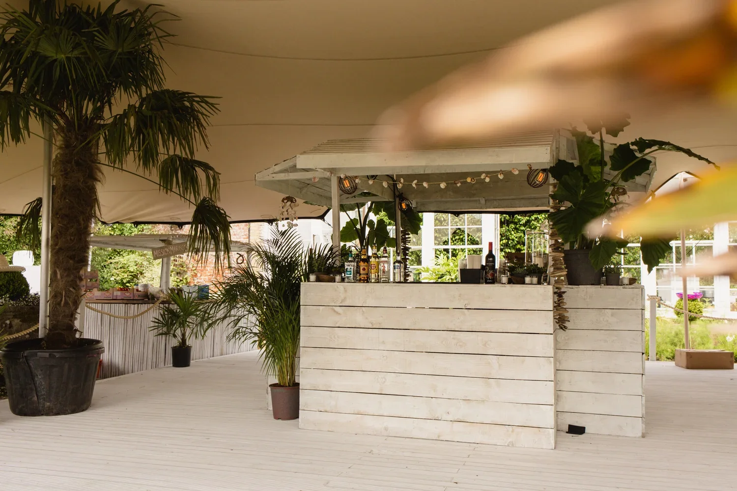 Outdoor bar with tropical plants, bar bottles, and string lights under a canopy in a garden setting.