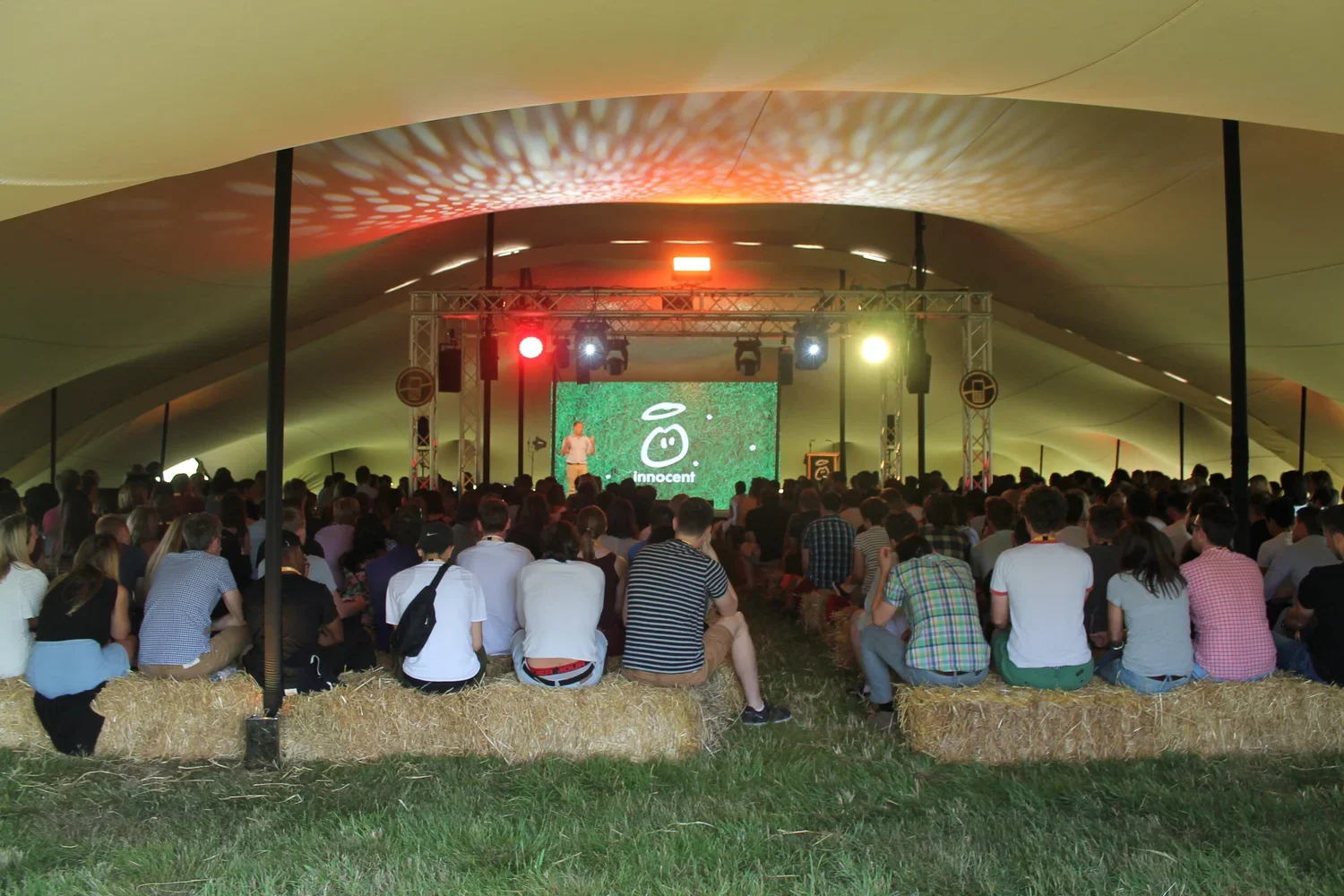 A large group of people seated on hay bales inside a tent, watching a stage performance with colorful lights and a large screen displaying 'innocent' and a logo.