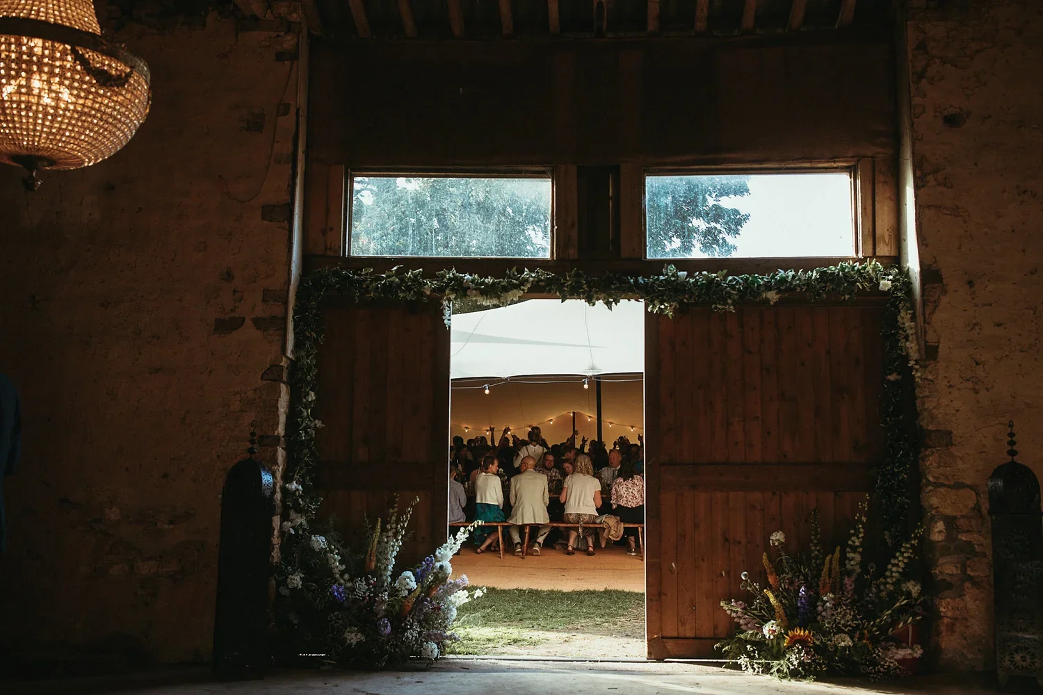 Open wooden barn door decorated with flowers, leading to a gathering of people in a tent with string lights inside.