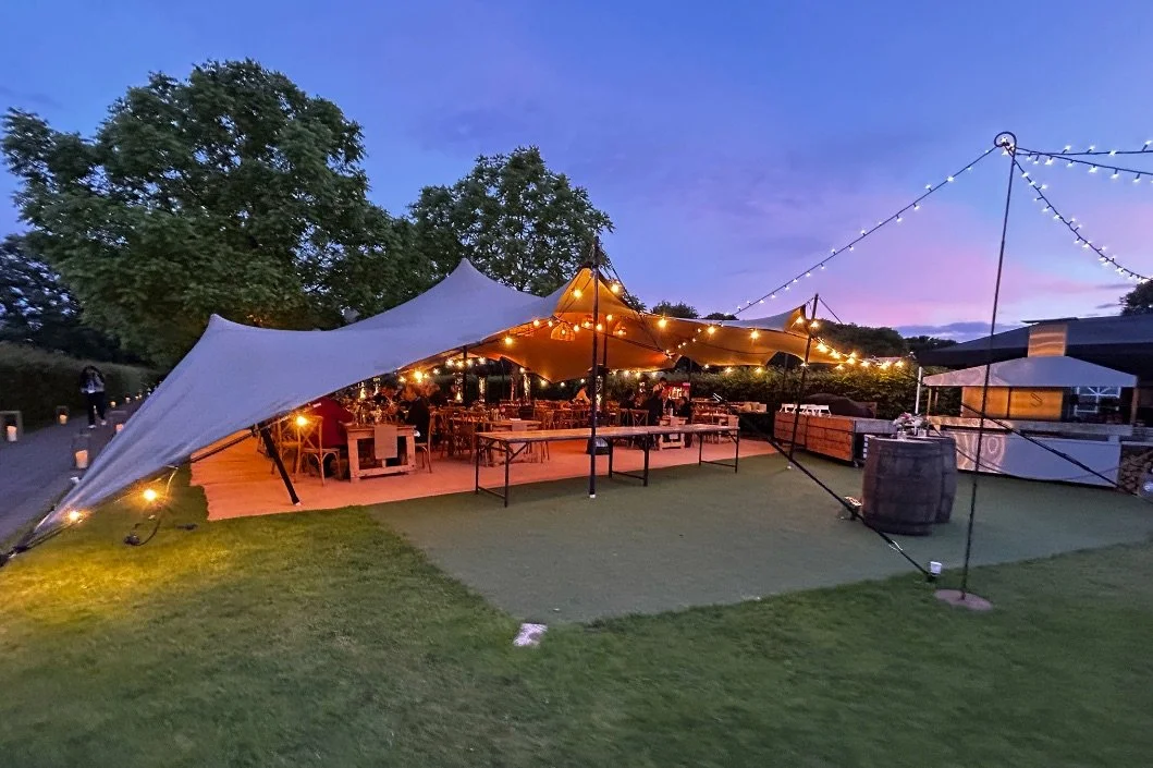 Outdoor evening event setup with string lights, a large canopy tent, tables and chairs, a barrel, and a bar area under a clear sky
