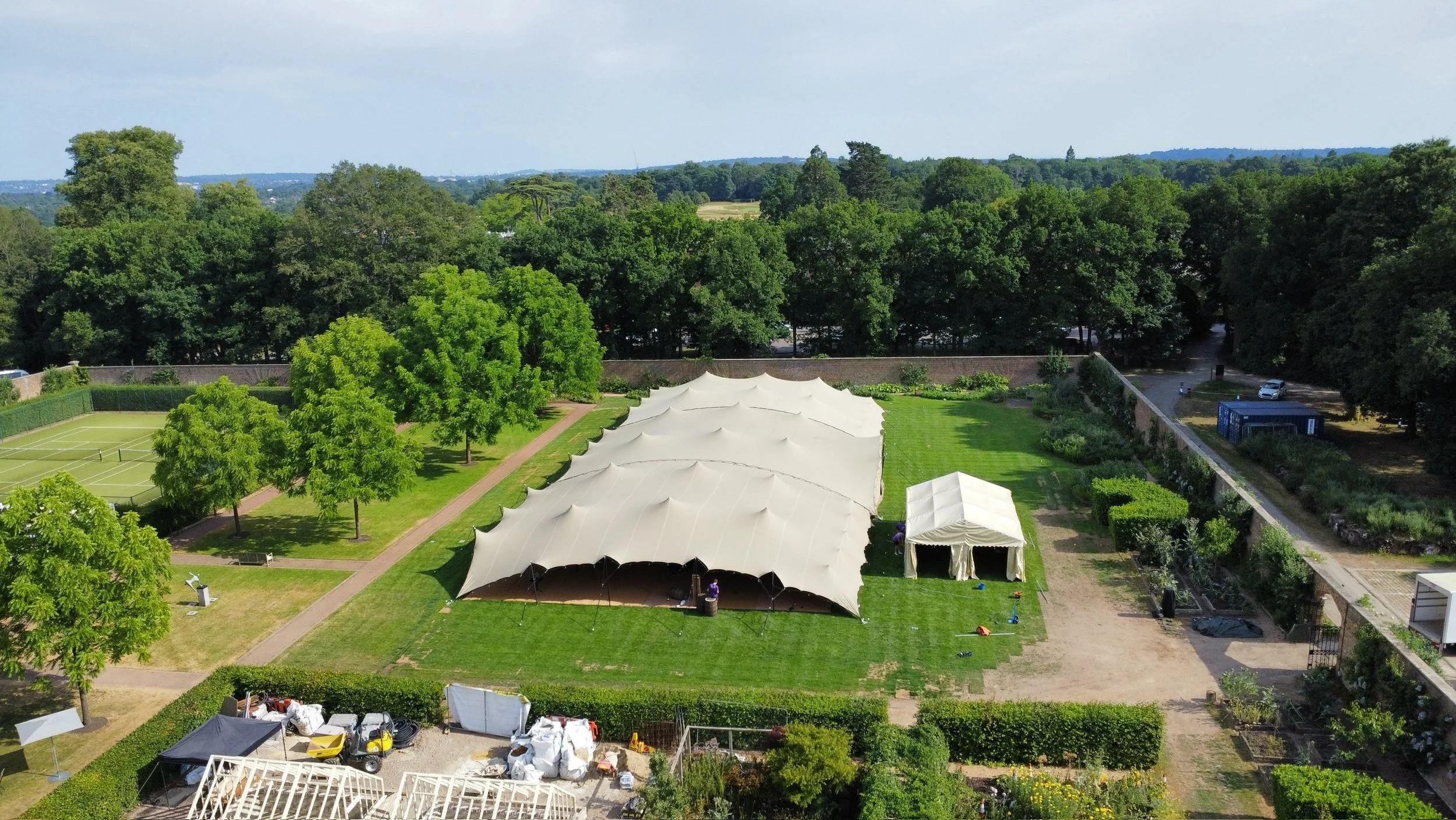 Aerial view of a garden with tennis courts, large white event tents, trees, and surrounding greenery.