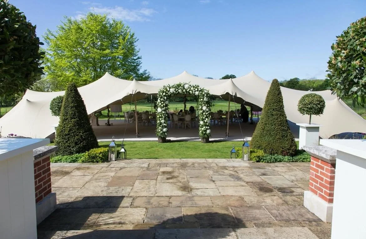 Outdoor wedding setup with a white tent, floral arch, and decorated tables at a garden venue.