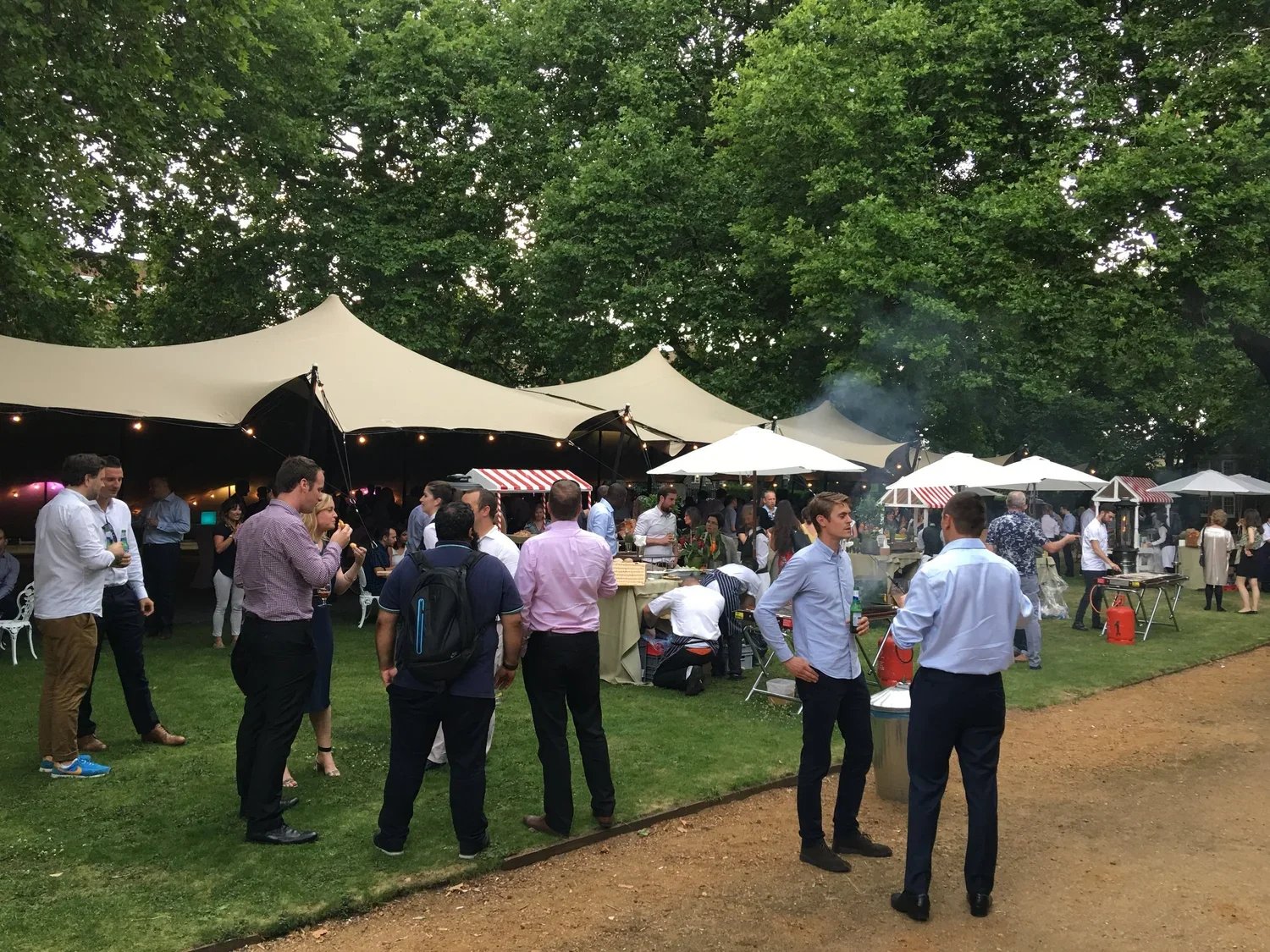 Outdoor event with people socializing under large white tents and umbrellas, surrounded by green trees.