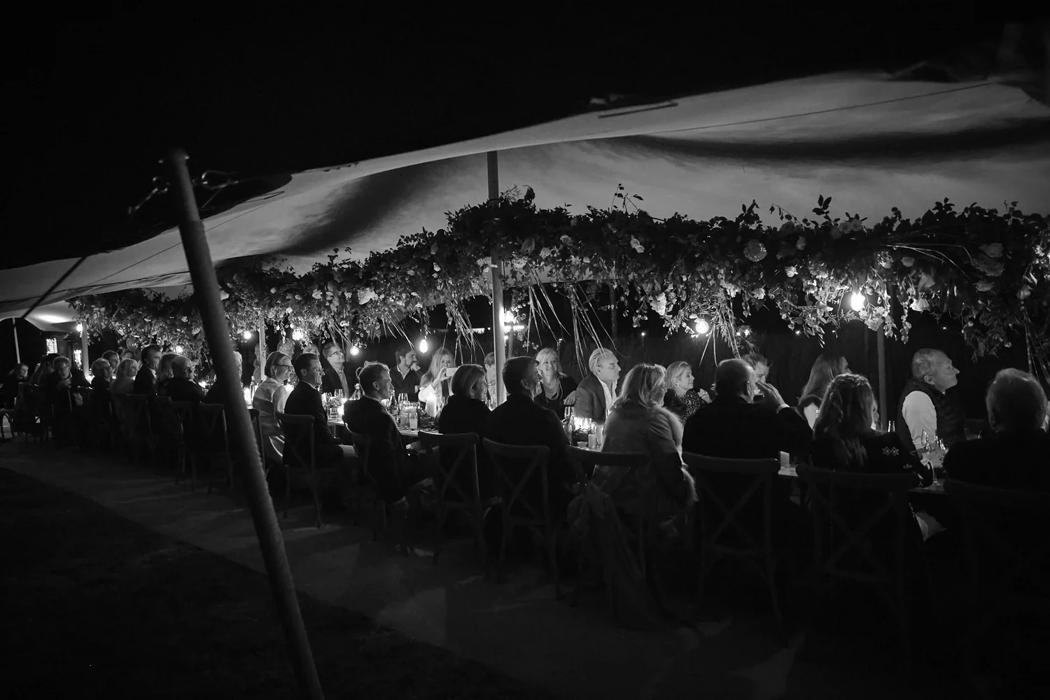 People seated at long tables outdoors during a nighttime event under a canopy decorated with flowers and string lights, with some tables lit by candles