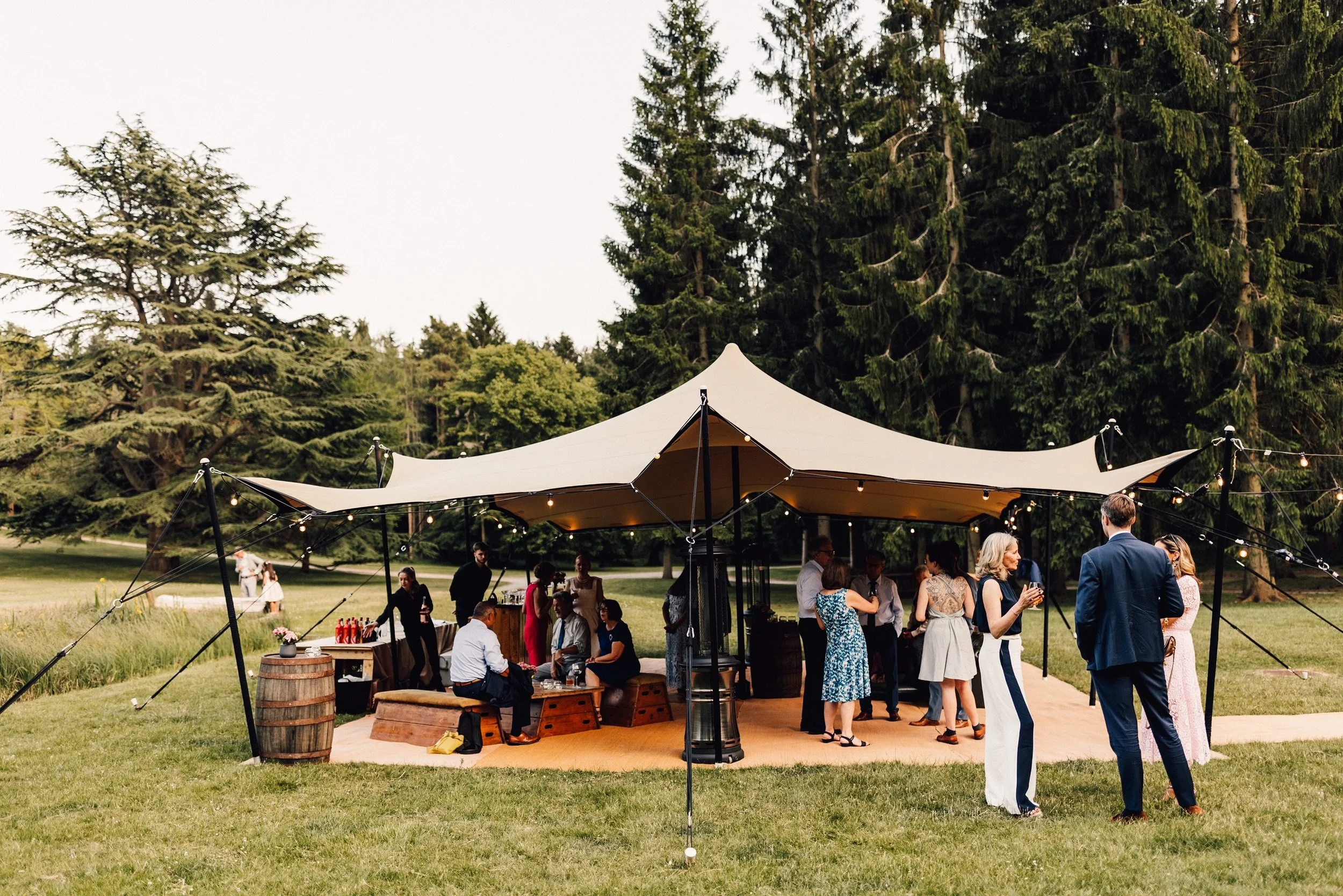 Outdoor gathering of people under a large beige tent with string lights, surrounded by tall green trees.