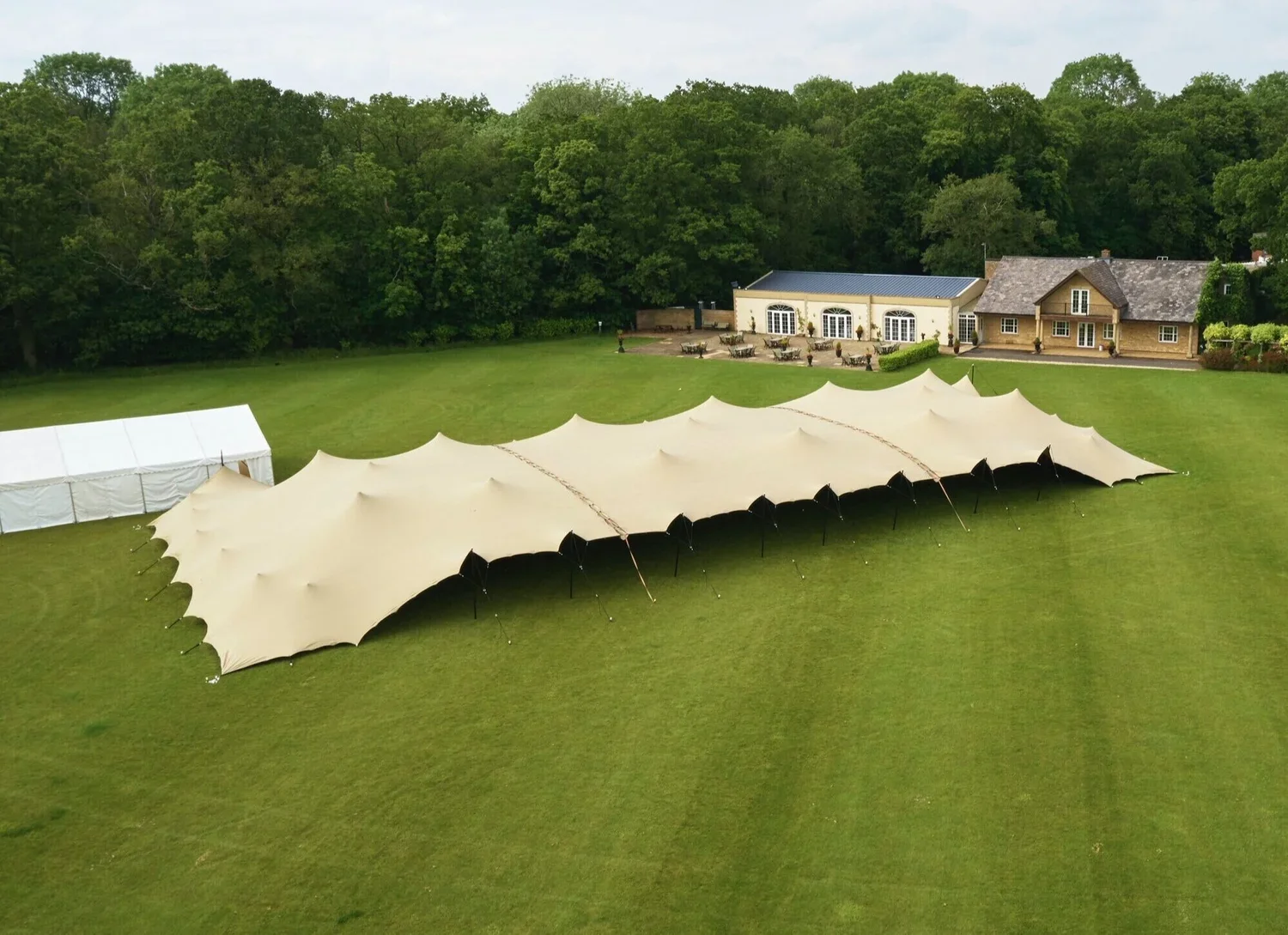Large outdoor event tent with multiple connected beige sections on a grassy field, with a white side tent nearby and a large house with patio and trees in the background.